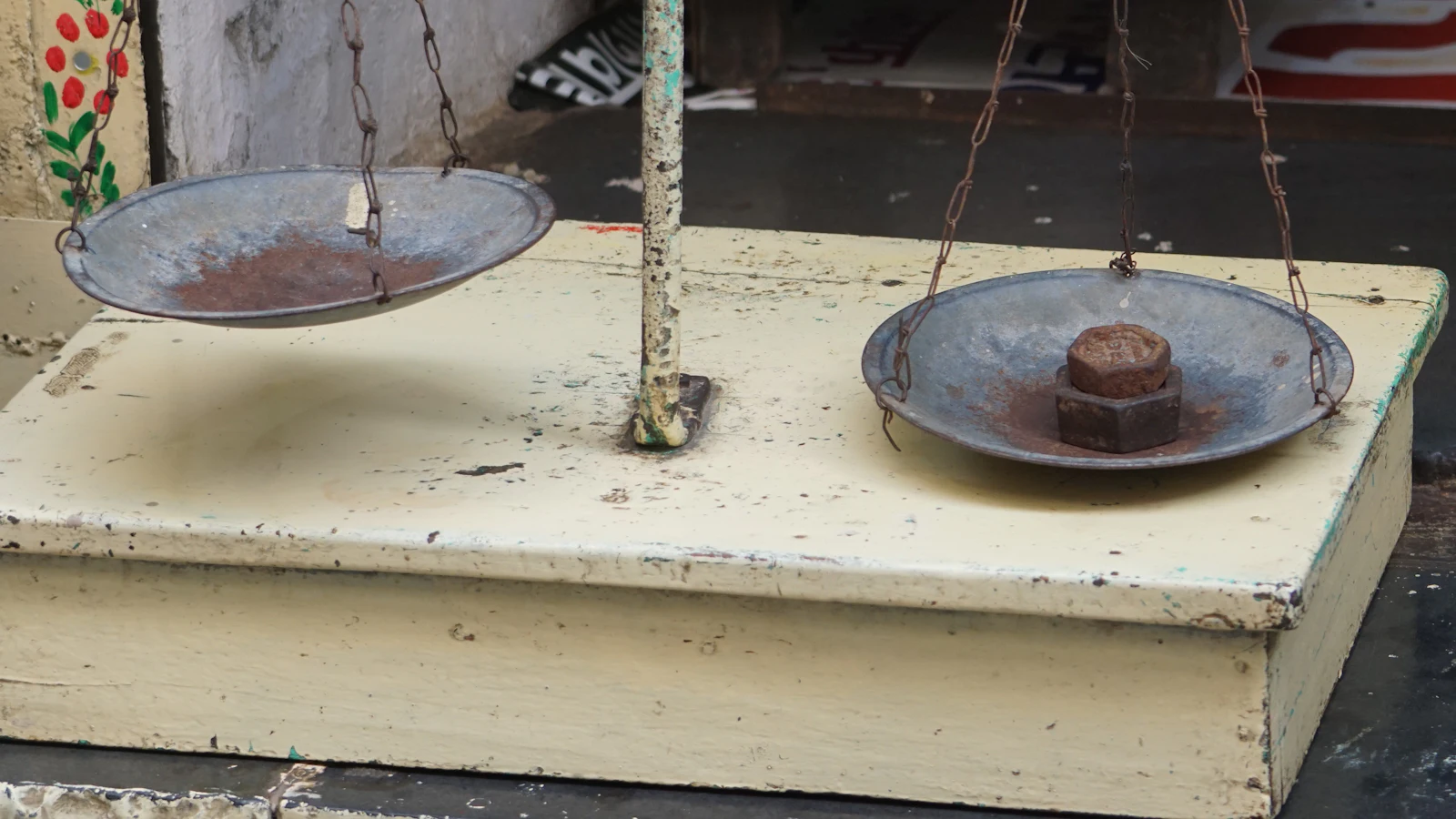A two-pan balance on a weathered counter, one pan empty and the other holding a rusted nut standing in for a calibrated weight — sizing against whatever reference happened to be to hand
