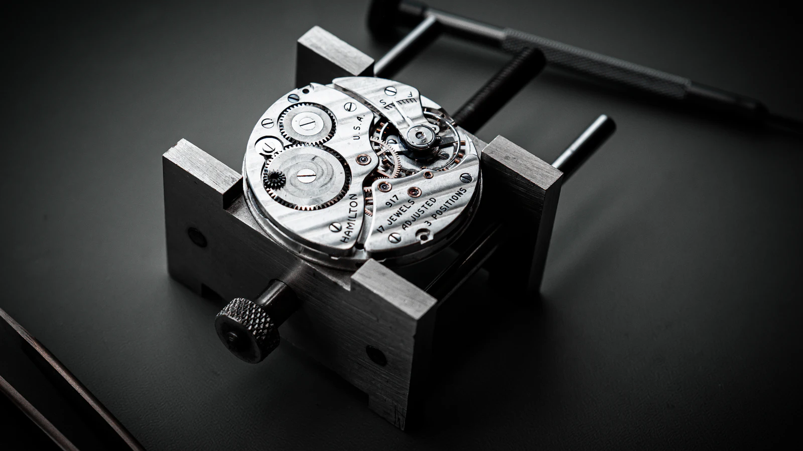 A pocket watch movement clamped in a steel movement holder beside a screwdriver: the fault held still on the bench so it can be examined, not described