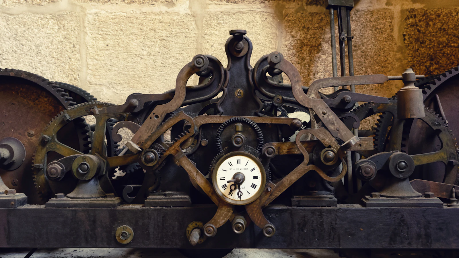 A tower clock movement on its iron bed against bare stone, gear trains and levers arranged around one small enamel dial — a large mechanism whose only job is cutting time into fixed intervals