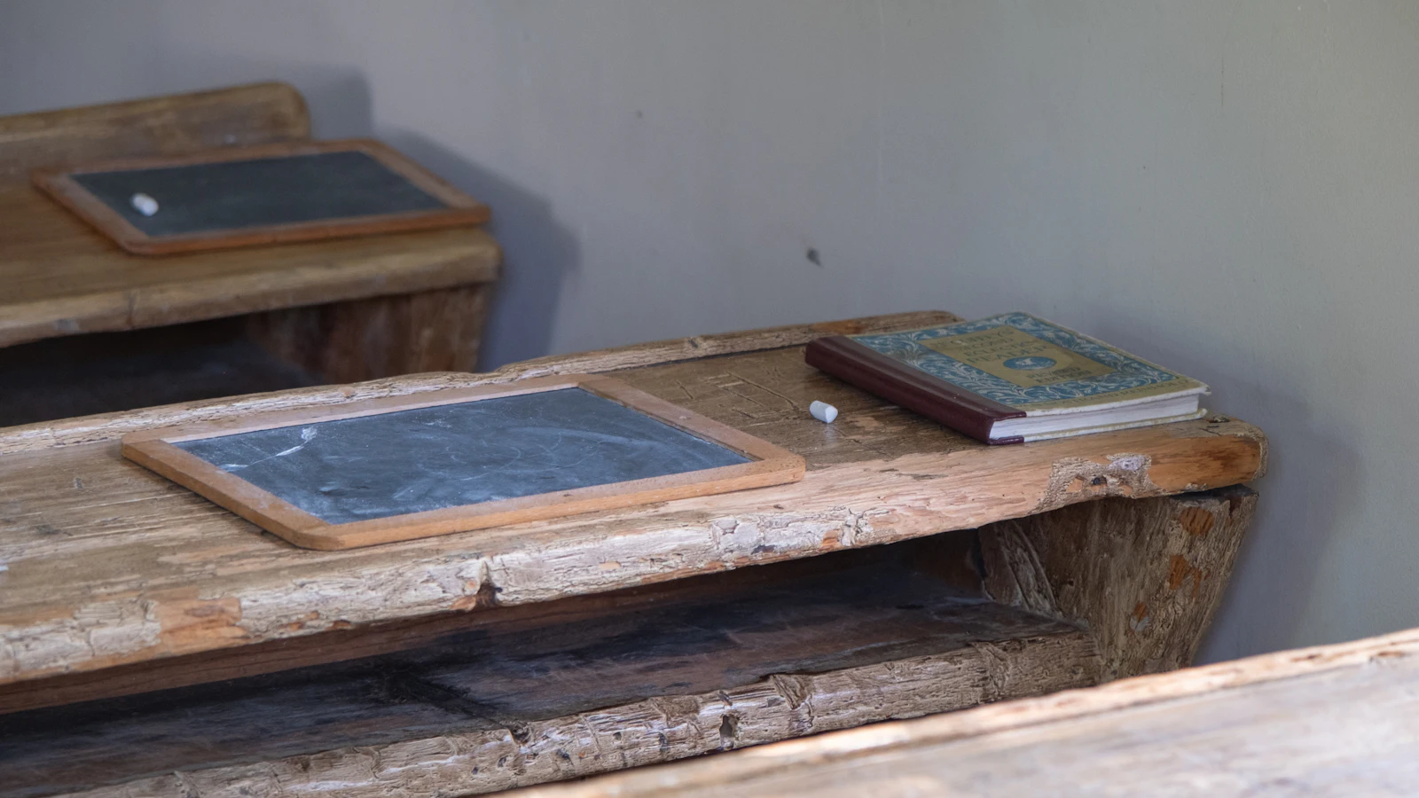Worn wooden school desks with a slate and a stick of chalk on each, the kind of surface where work is written, checked, wiped and written again until it comes out right